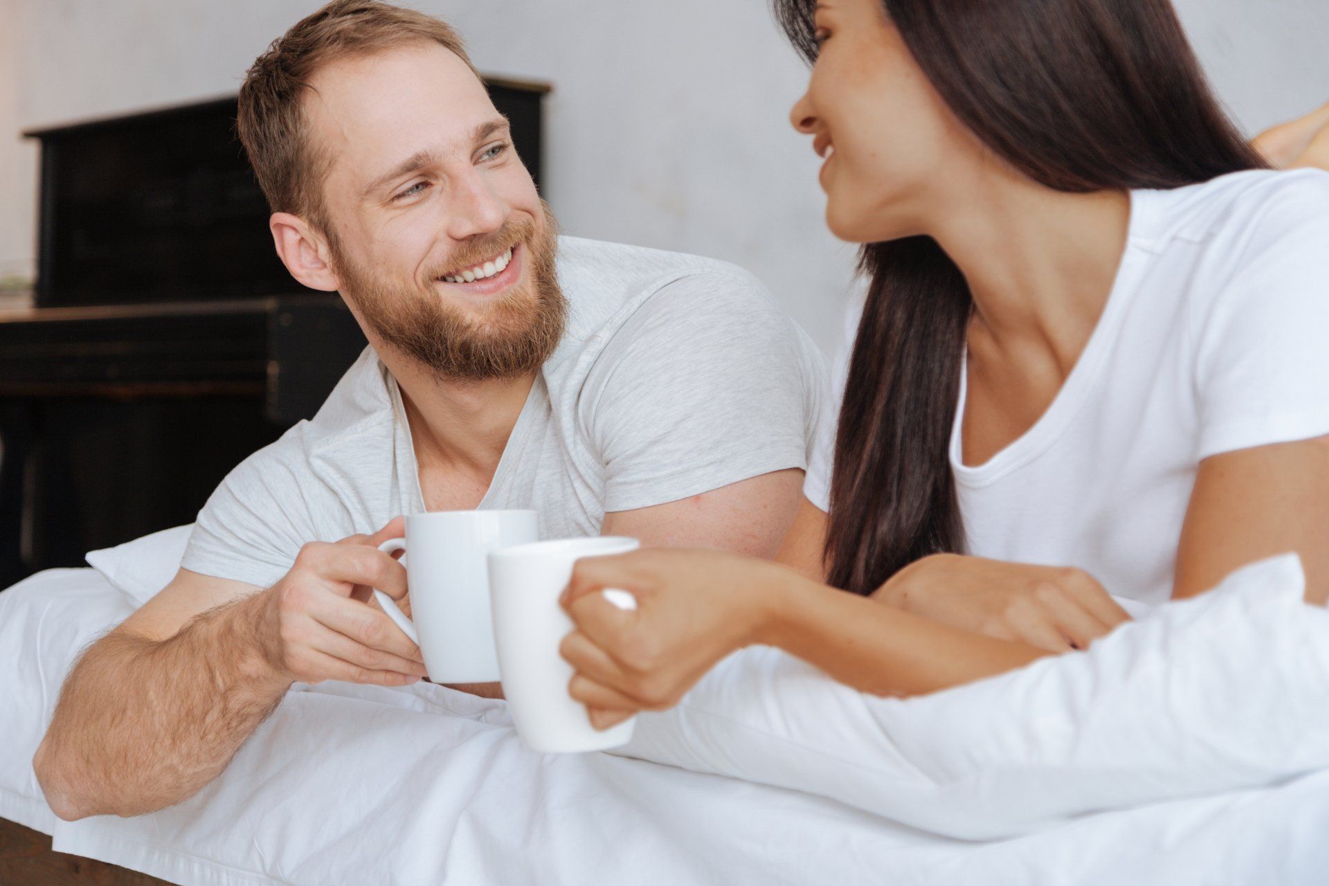 A man and a woman are sitting on a bed drinking coffee.