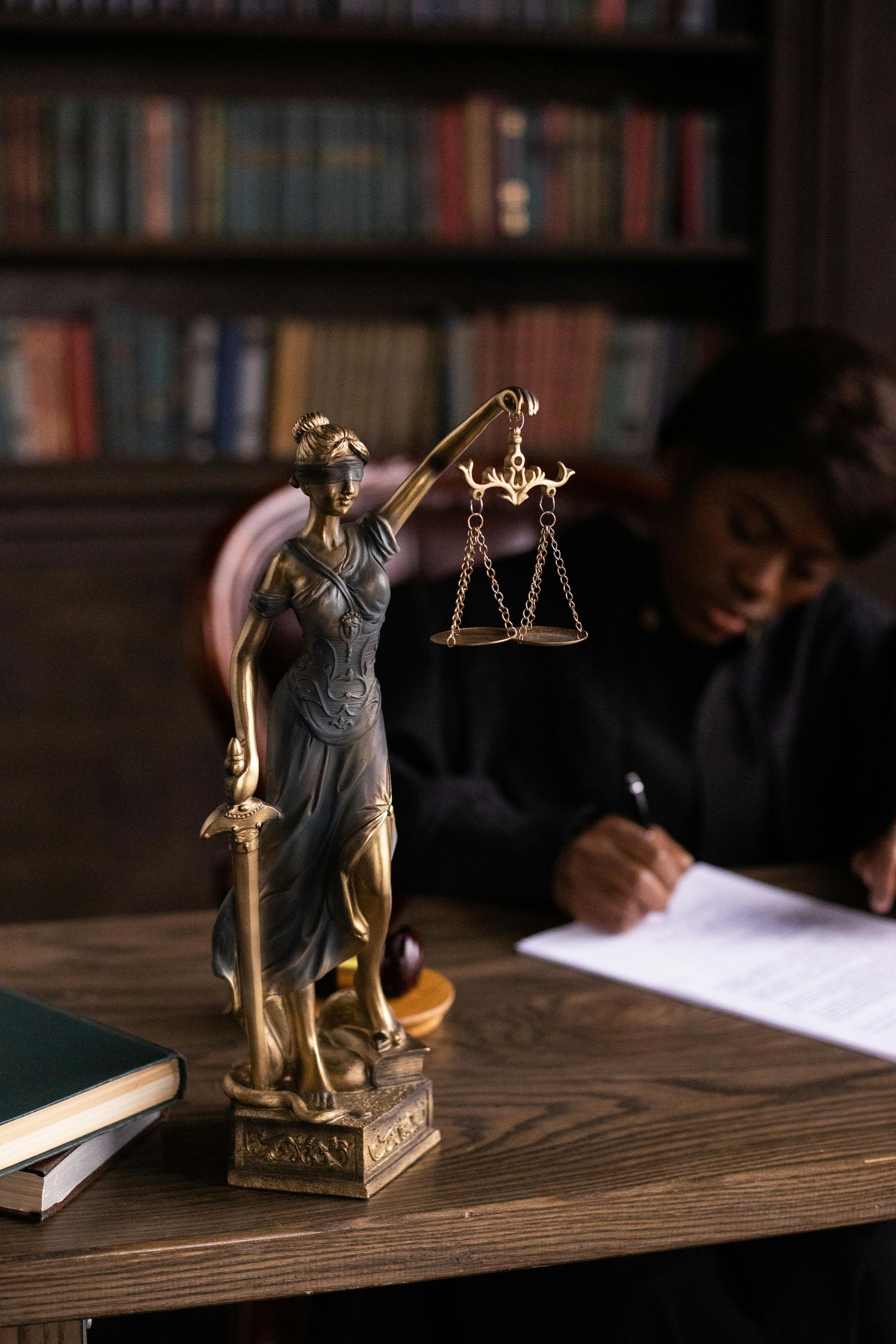 A statue of justice is sitting on a wooden table next to a man writing on a piece of paper.