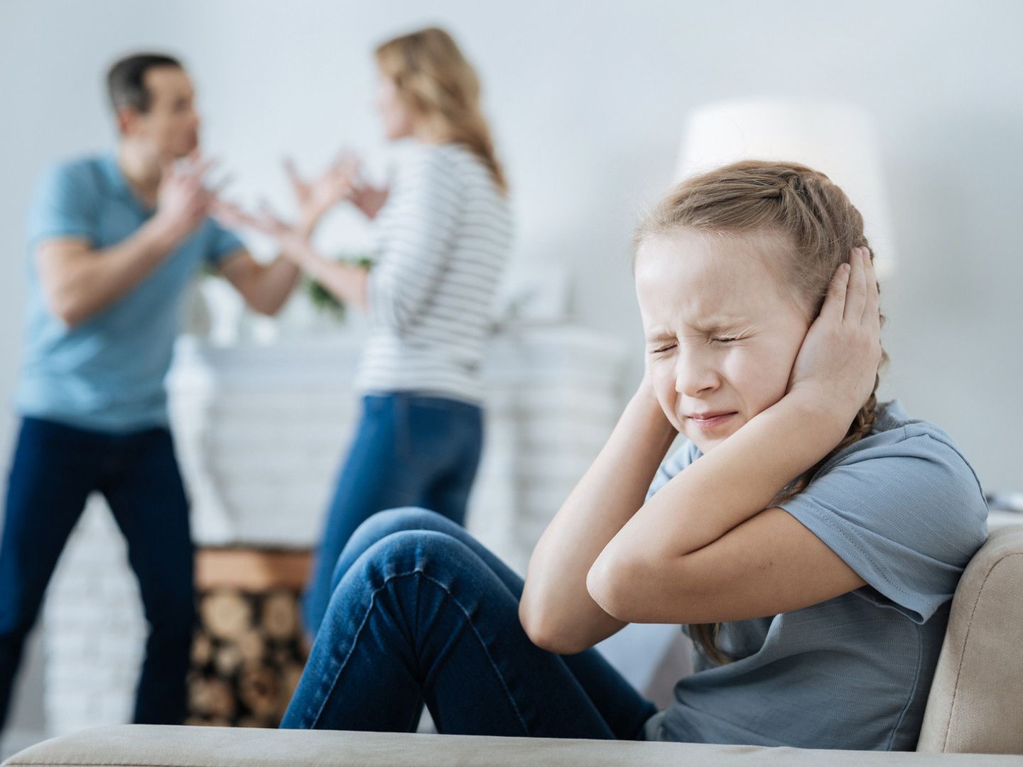 A boy is sitting on a couch covering his ears while his parents argue in the background.