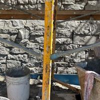 Yellow rusty metal post with crossed supports in front of a stone wall and bucket