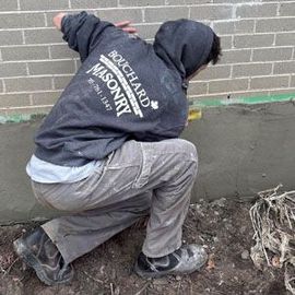 Person crouching by a brick wall, wearing a black hoodie and gray pants, working in dirt near exposed roots.