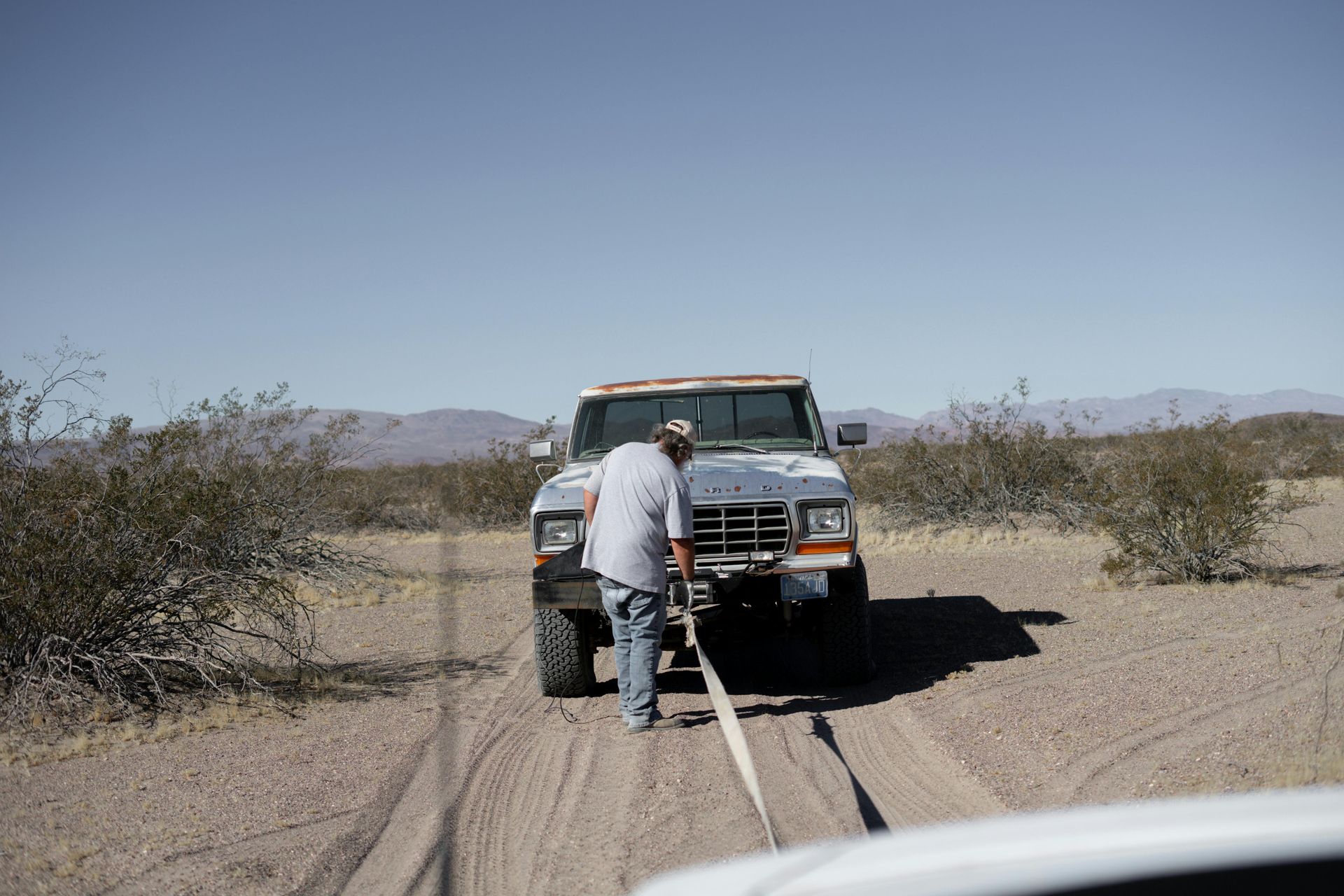 Man pulling a tow strap to a white truck on a dirt road in a desert setting.