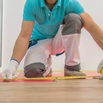 Person kneeling on a hardwood floor measuring with a tape measure and pencil, wearing a teal shirt and work gloves
