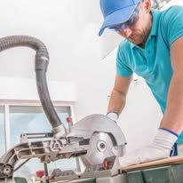 Worker using a circular saw on a workbench, wearing blue cap, shirt, and gloves in a workshop.