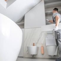 Person in gray installs a wall-mounted sink in a modern bathroom with white fixtures.