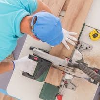 Worker using a power saw to cut wood on a workbench, wearing a blue cap and gloves.