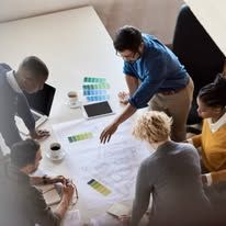 Team meeting around a table, pointing at printed charts and a tablet in a bright office.