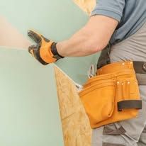Person in work gloves fastening a wooden panel to a wall, with a tool belt and gray shirt.