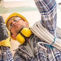 Worker in hard hat and safety glasses reaching overhead with coiled cable in a construction setting