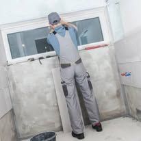 Person taking a mirror selfie in a white room, wearing a gray outfit and cap.