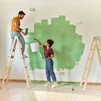 Two people painting a wall green on wooden ladders in a bright room