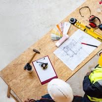Top-down view of a workbench with a blueprint, yellow tape measure, tools, and a hard hat on plywood.