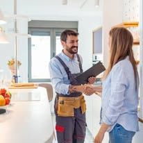 Two people in a bright kitchen shake hands, one holding a clipboard and wearing a tool belt.
