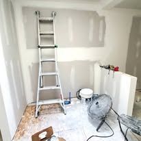 Interior room under renovation with ladder, paint bucket, fan, and unfinished drywall patches