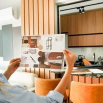 Person holding a floor plan in a modern living room with orange chairs and a kitchen in the background