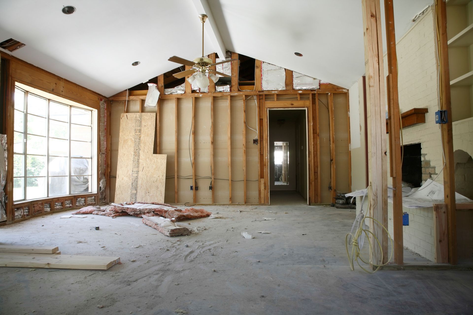 Unfinished room under renovation with exposed wall studs, debris on the floor, and large windows.