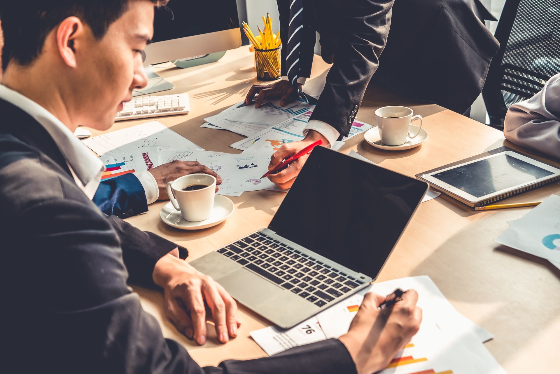 People in a meeting around a laptop, reviewing documents and coffee on a bright conference table.