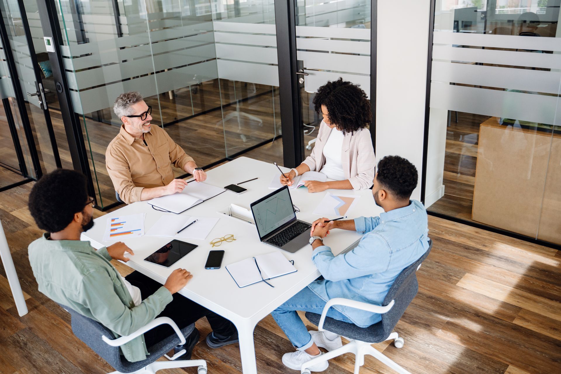 Four coworkers meeting around a table in a glass-walled office, discussing papers and a laptop.
