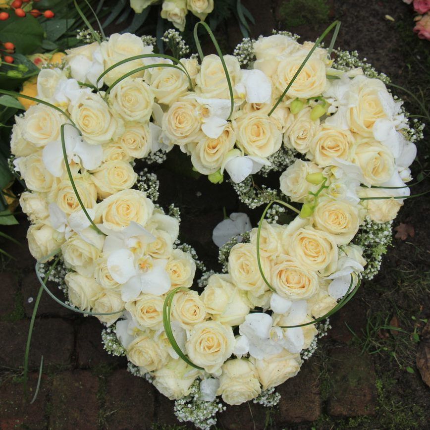 A heart-shaped floral wreath made of pale yellow roses, white orchids, and baby's breath on a dark brick surface.