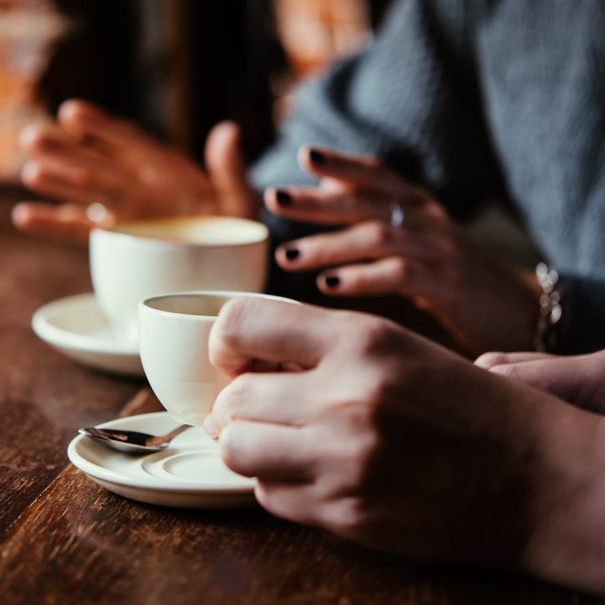 Two people at a wooden table engage in conversation over cups of coffee, one gesturing while the other holds a cup.