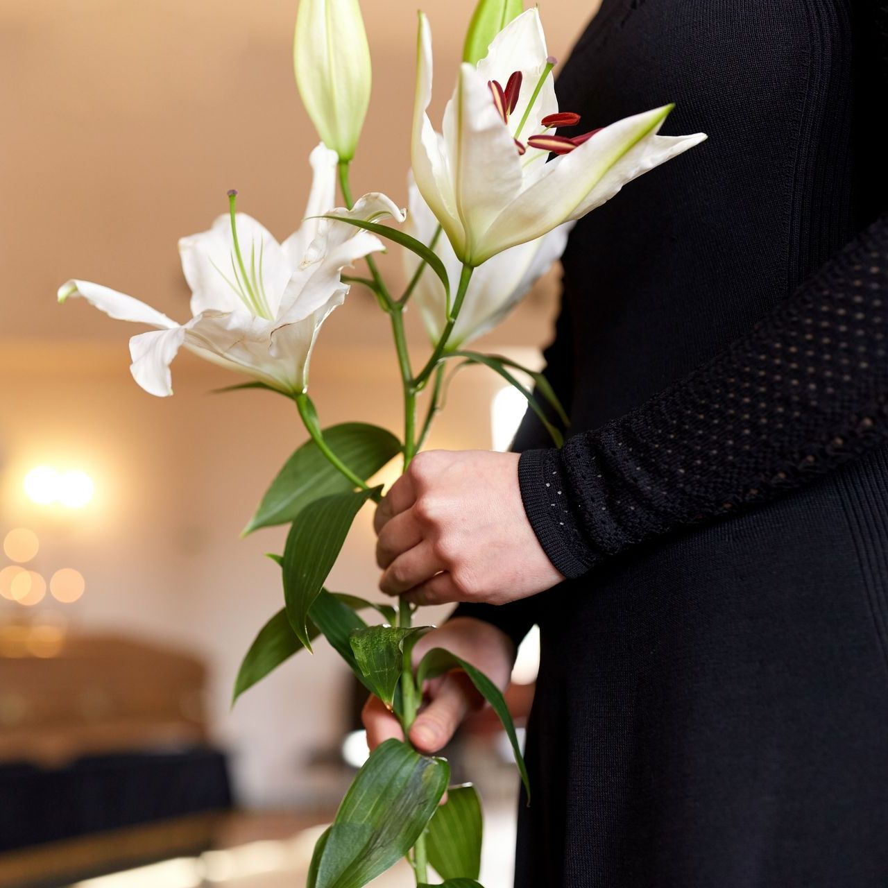 A person in a black long-sleeved dress holds a bunch of white lilies against a softly lit indoor background.