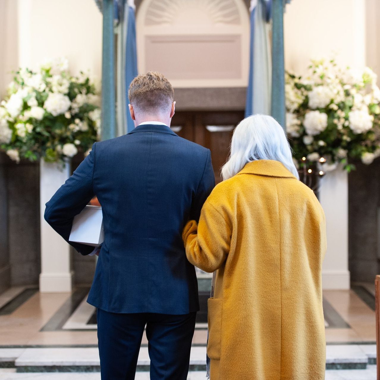 A person in a dark suit and a person in a long yellow coat stand before a doorway flanked by large white floral bouquets.