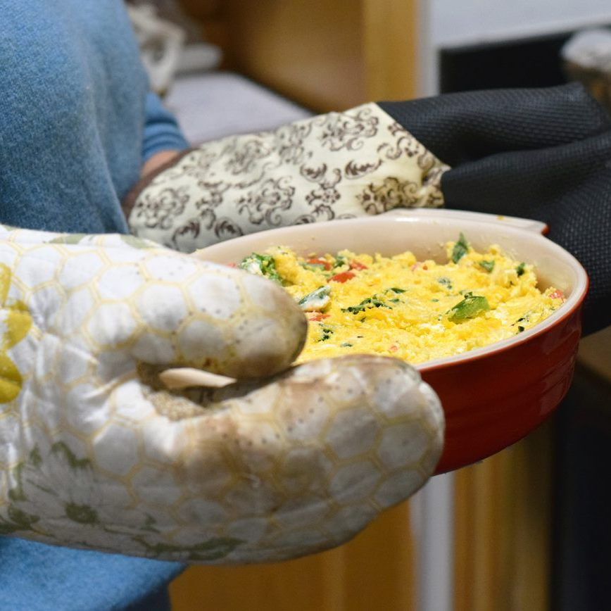 A person wearing oven mitts holds a red ceramic baking dish filled with a baked egg and vegetable dish.