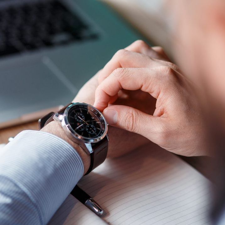 A person in a light blue shirt adjusts the crown on a round, black-faced chronograph watch while working at a desk.