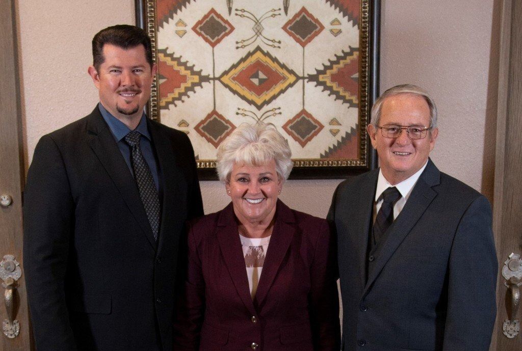 Three people stand smiling in suits before a wall decorated with a geometric-patterned framed artwork.