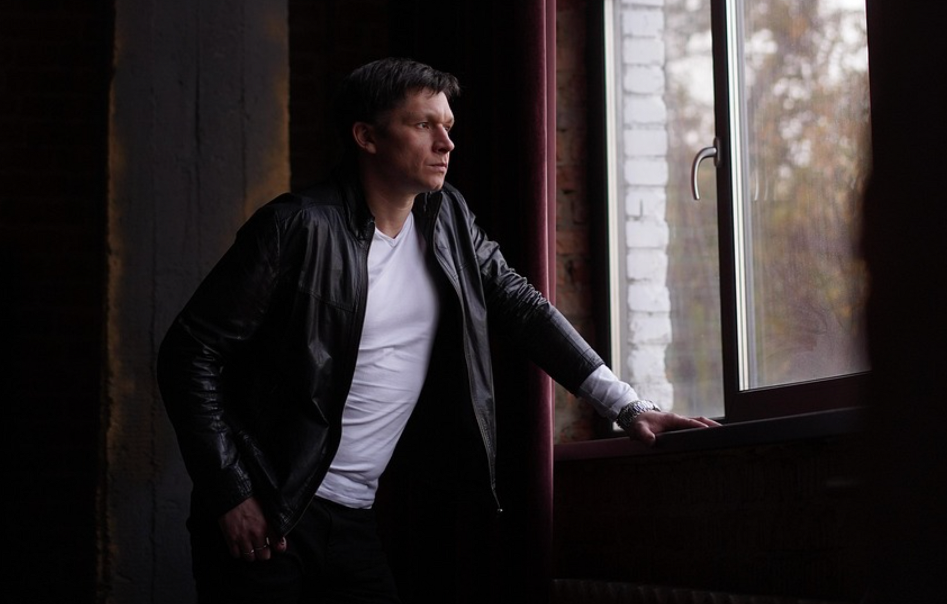 A man in a leather jacket is standing in front of a window concerned about being let go