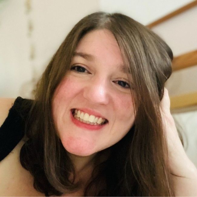 Woman with long brown hair smiling, resting head on hand, in a lit setting.