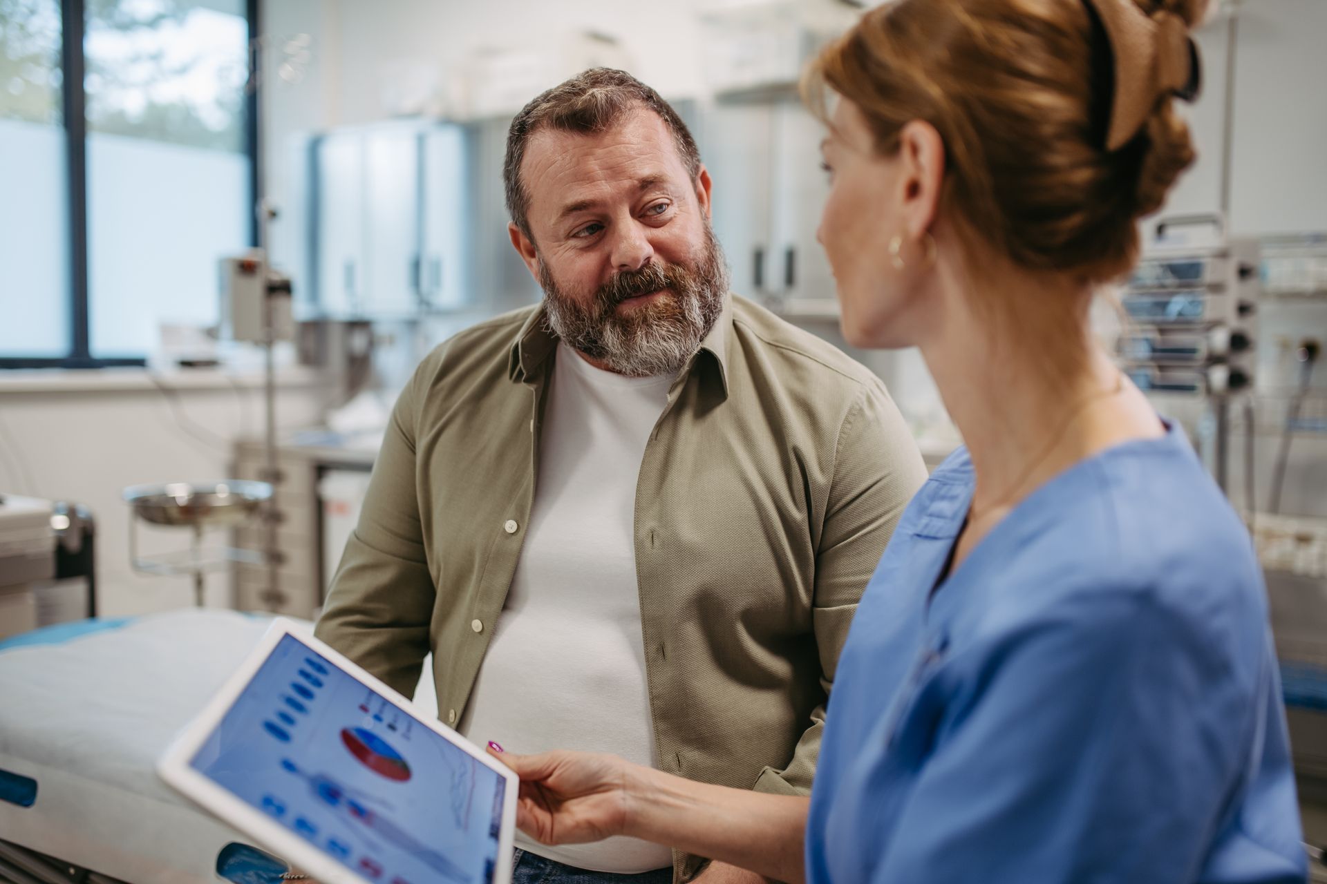 Clinician in scrubs reviews a tablet health chart with a male patient in a medical office