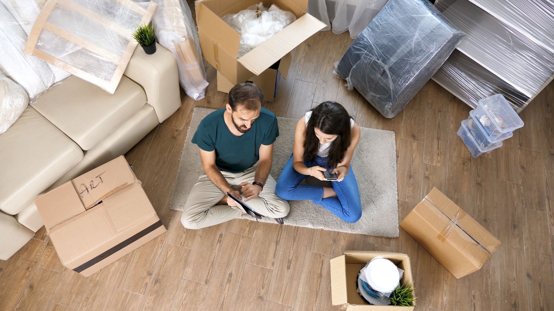 A man and a woman are sitting on the floor in a living room surrounded by cardboard boxes.