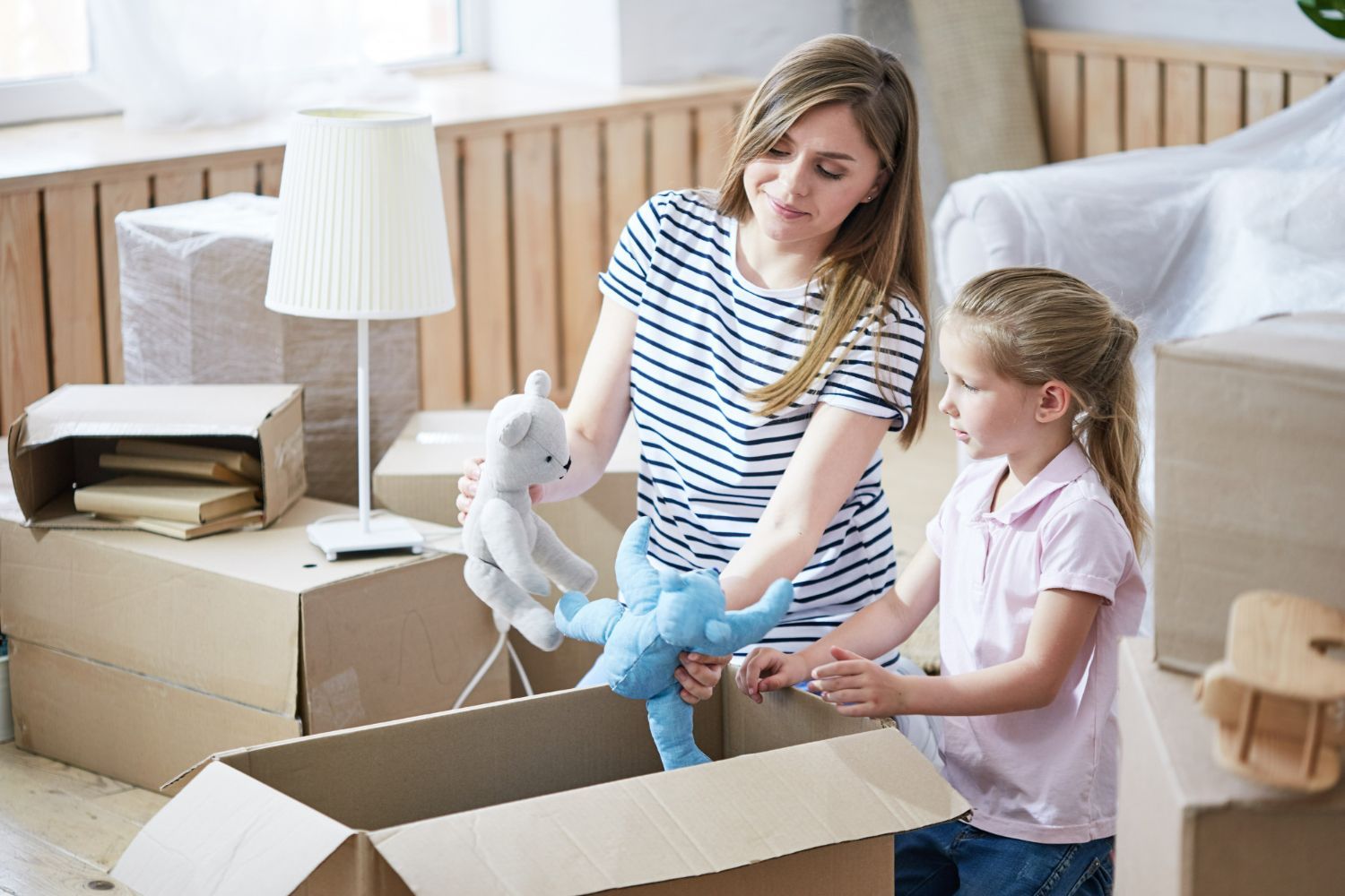 Woman and girl unpack stuffed animals from a cardboard box in a bright bedroom