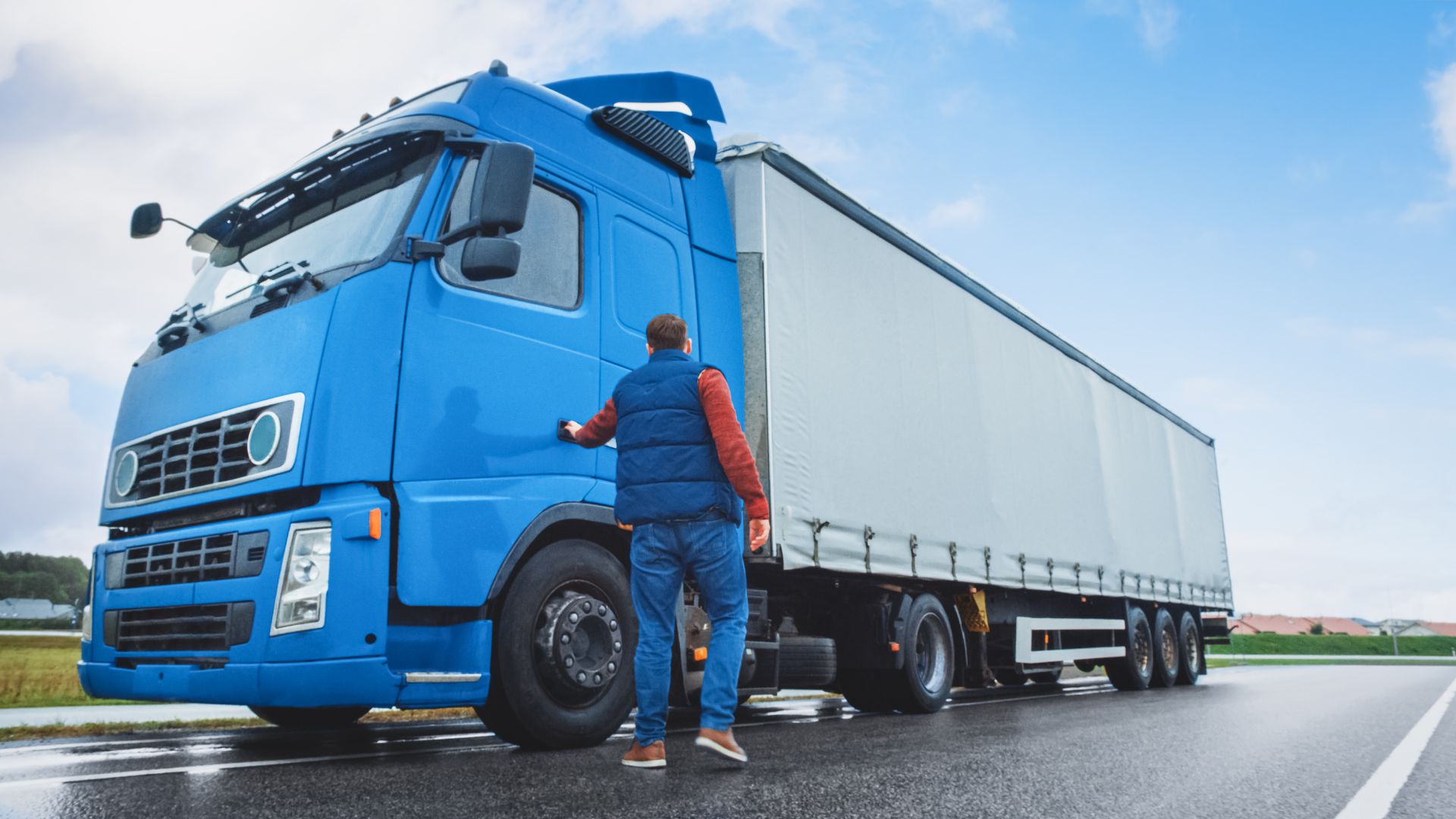Blue semi-truck on road, driver in vest opening door. Cloudy sky.