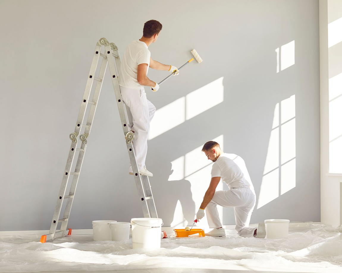 Two people painting a gray wall with paint rollers. One is on a ladder, the other on the floor.