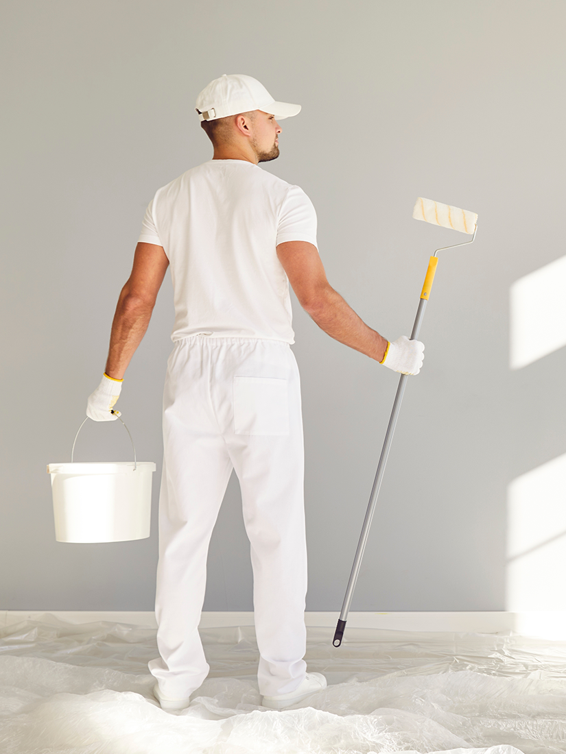 Person in white paint suit, holding a paint roller and bucket, facing a gray wall.