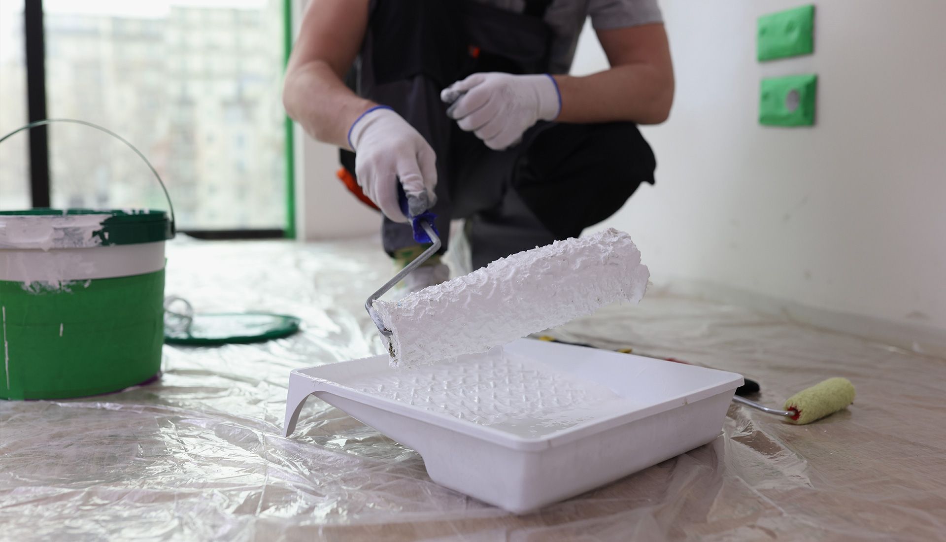Person in overalls and gloves paints wall with white paint using a roller and tray.