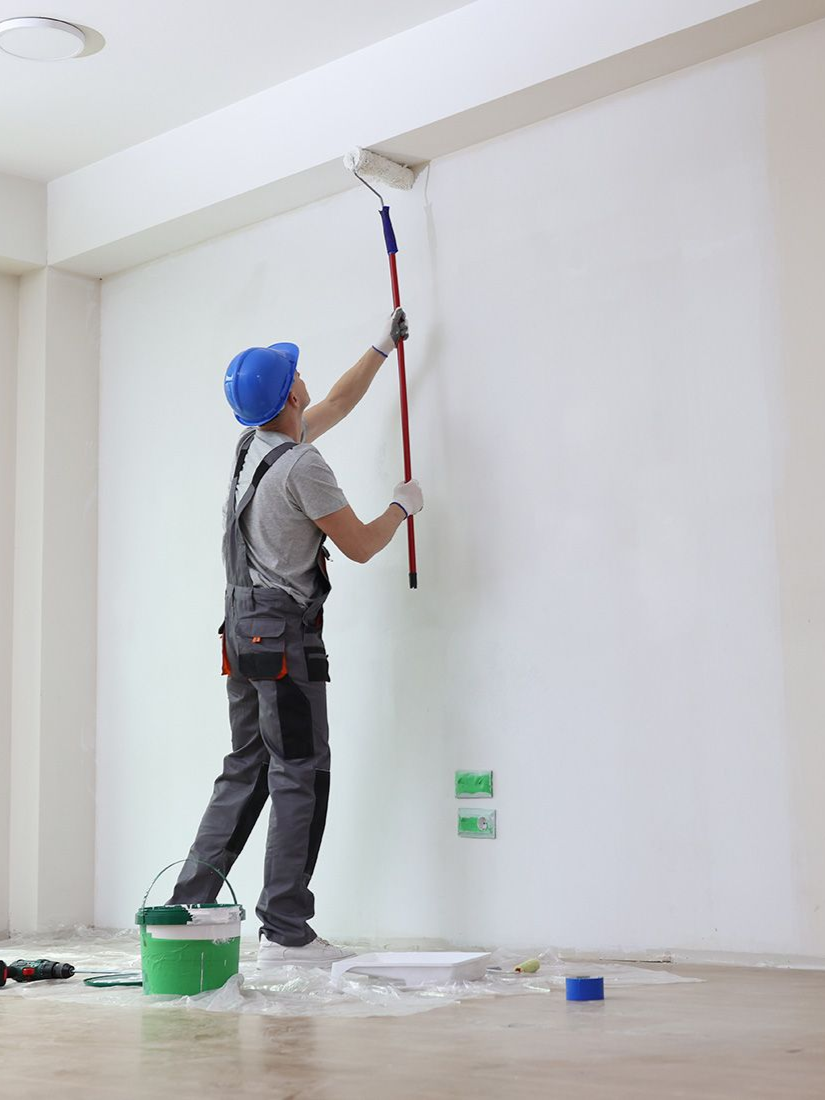 Person in overalls paints a white wall with a roller. There's a bucket of paint, a hard hat, and some tools.