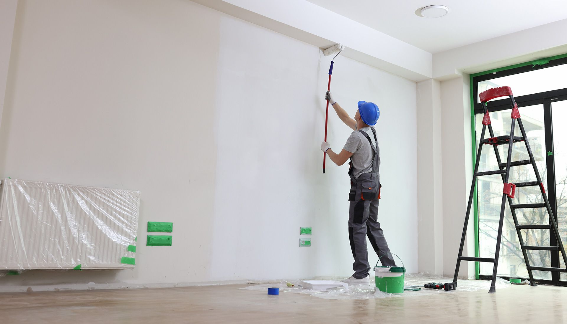 Person paints a wall white with a roller; a ladder, paint bucket, and taped-up heater are in the room.