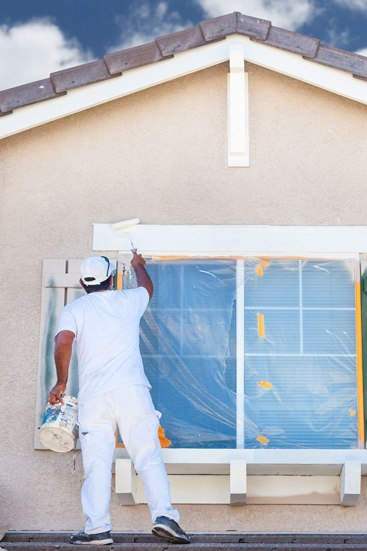 Person painting the trim of a house window, wearing white clothing, sunny day.