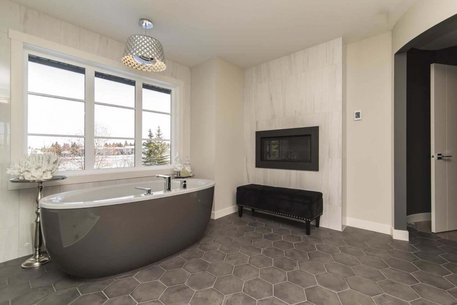 Spacious bathroom with oval bathtub, fireplace, and large window. Gray hexagonal floor tiles and white walls.