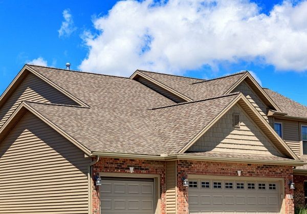 Tan roof with blue sky and clouds; beige siding, brick facade, and garage doors.