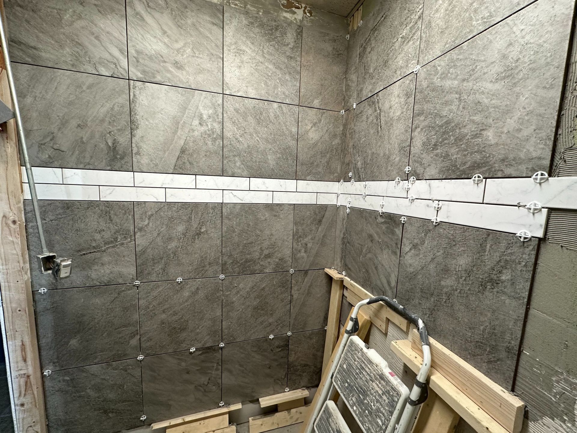Gray tiles being installed on bathroom walls with spacers. Ladder and wood supports present.