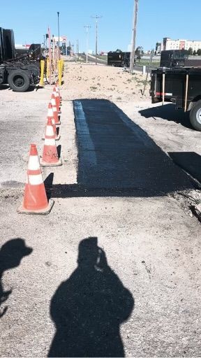 Freshly paved asphalt strip, orange cones line edge, person's shadow visible.