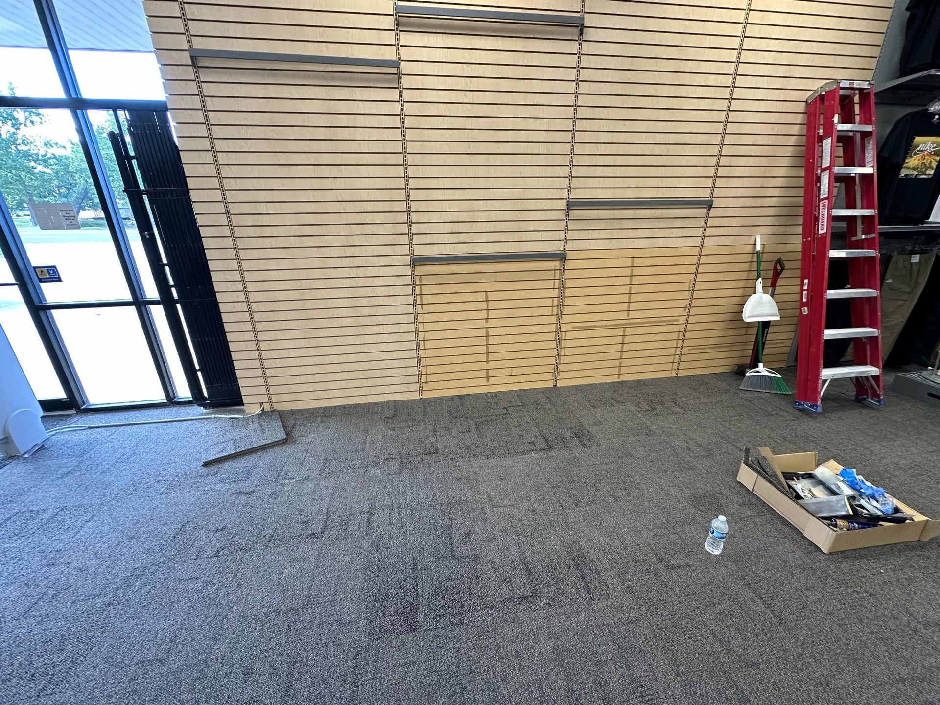 Room with a patterned wood wall, carpeted floor, ladder, and supplies; near a glass door.