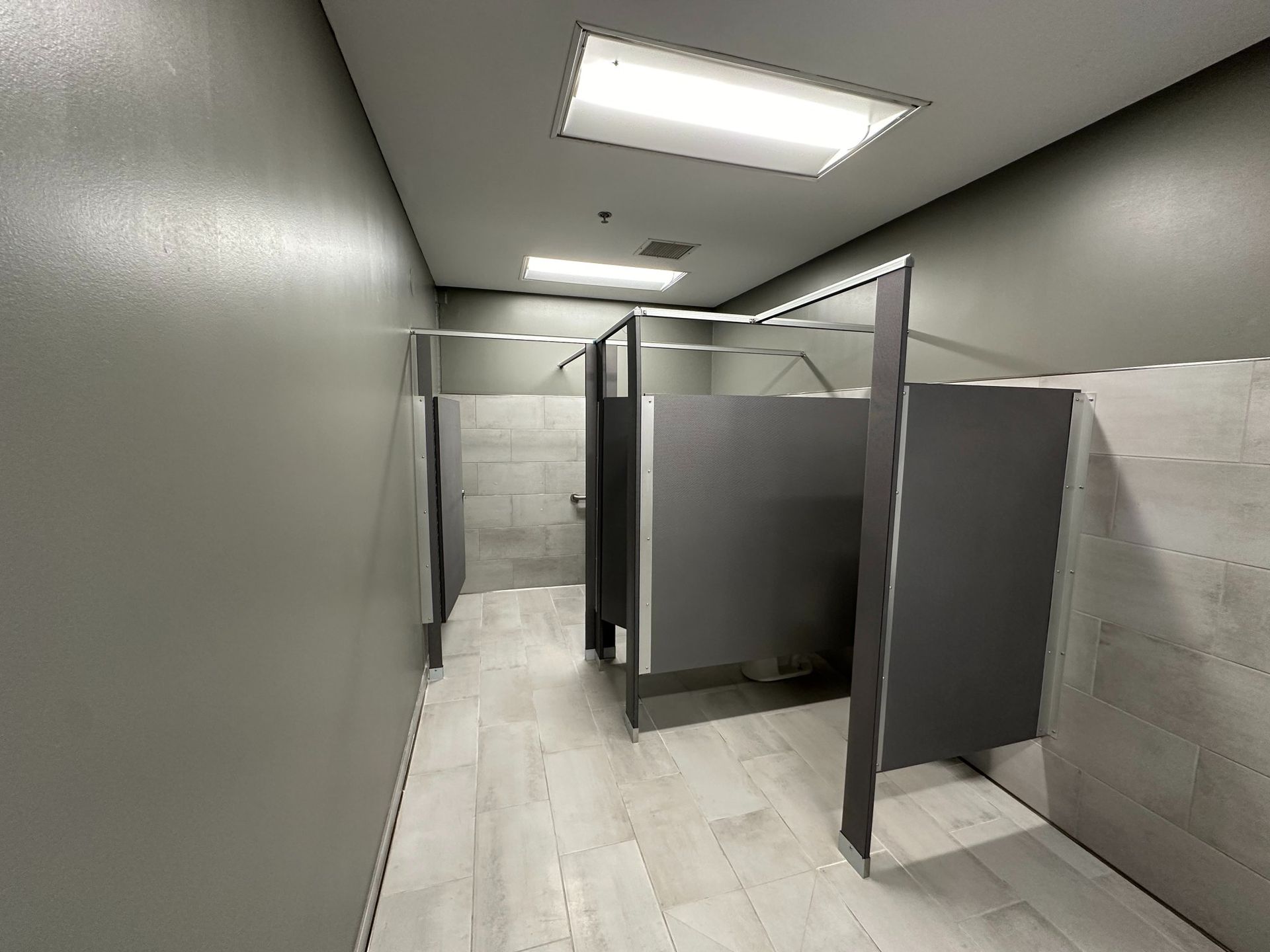Public restroom with gray stalls, light wood-look floor, and bright overhead lights.