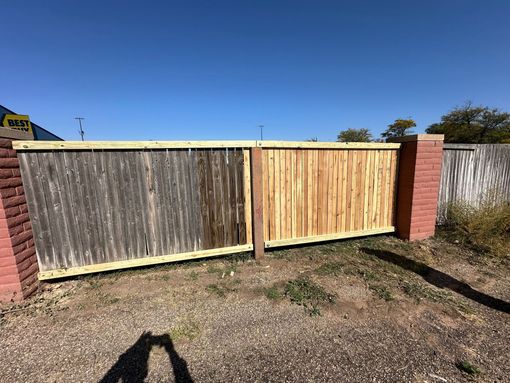 A wooden fence, half weathered grey and half new, flanked by brick columns, sits against a blue sky.