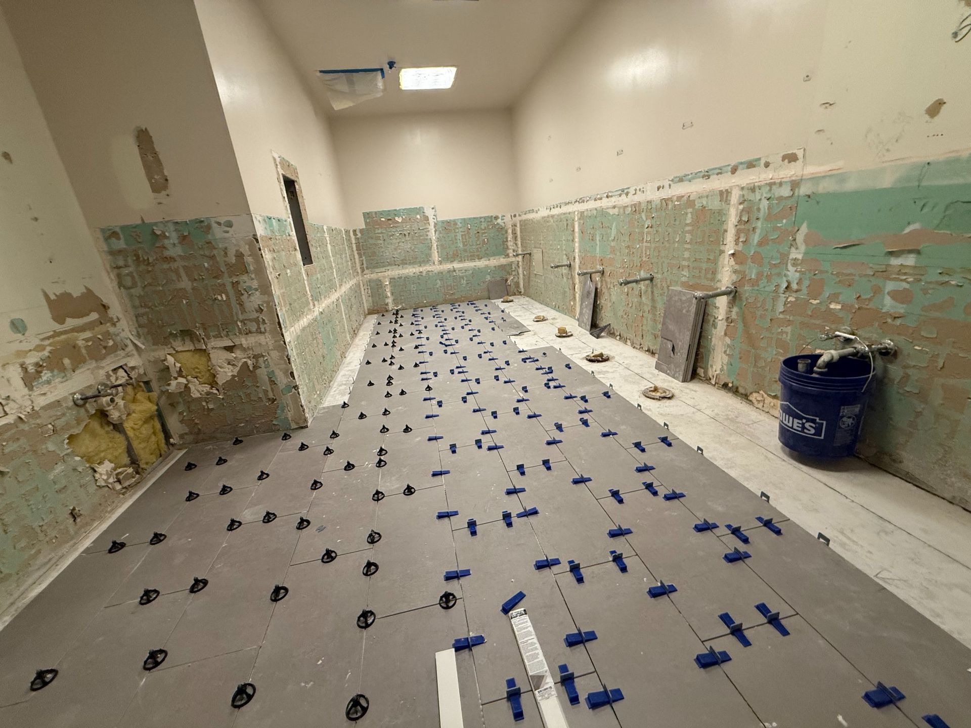 Room undergoing tiling. Gray flooring with blue leveling clips. Walls partially stripped with some tiles remaining.