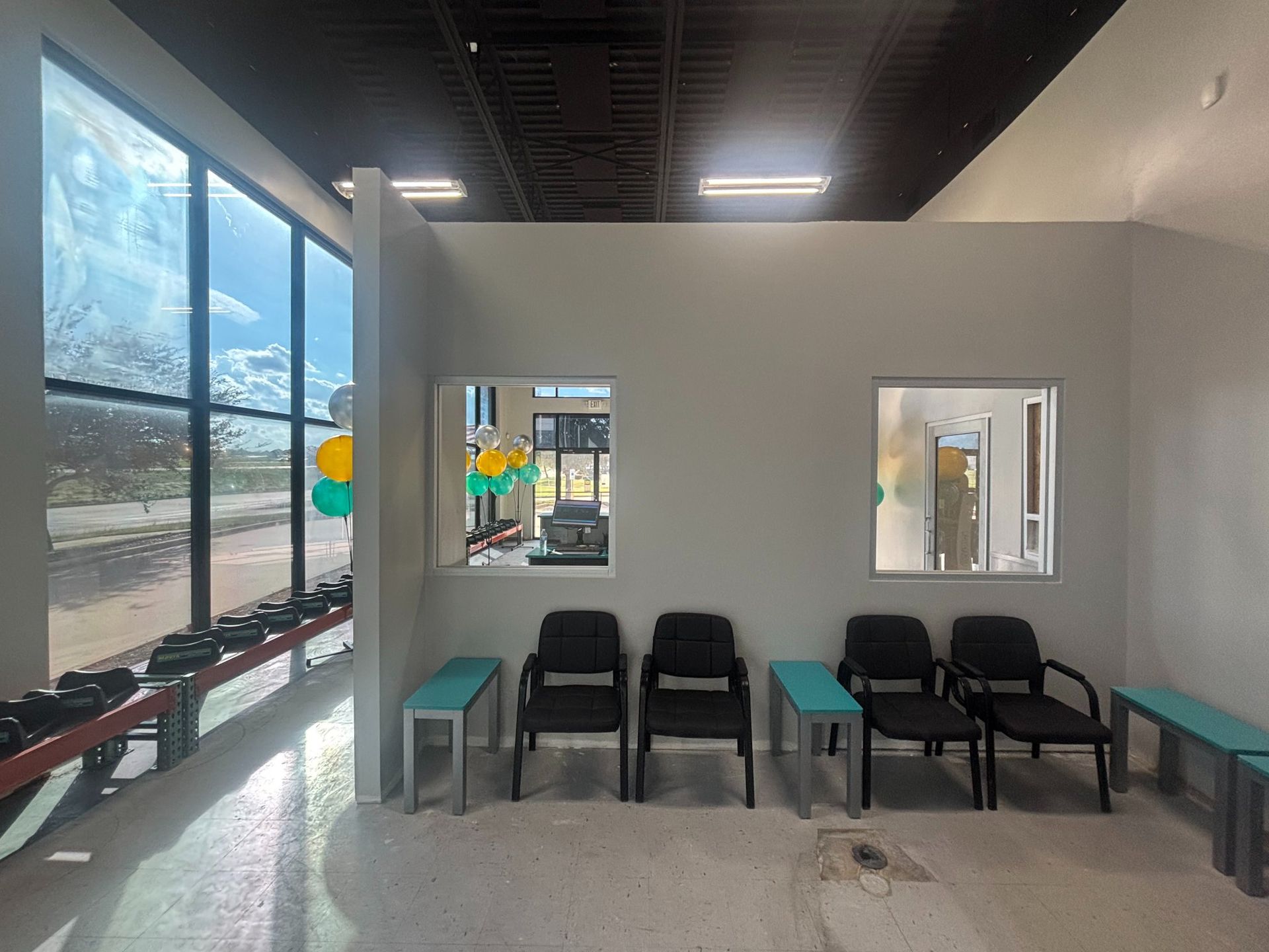 Interior of a waiting area with chairs, windows, and dumbbell rack; light gray walls, concrete floor, and large window.